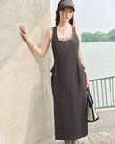 Woman strolling a weekend market in a sage-green pinch-pleat pocket dress, airy organic cotton and deep side pockets pair utility with feminine flow for hot-summer comfort.